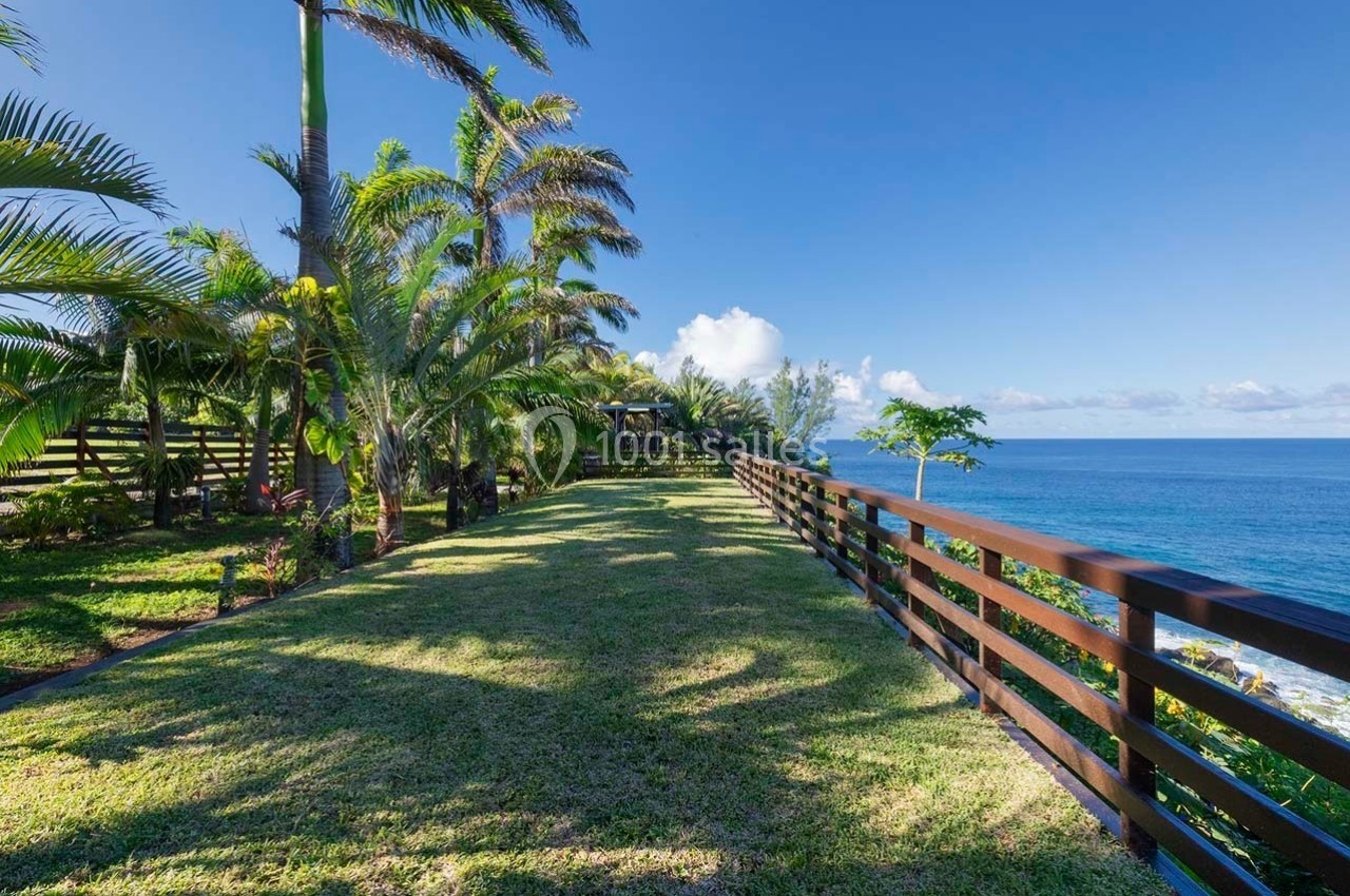 Jardin bordé de palmiers avec une pelouse verte, une clôture en bois et vue sur l'océan sous un ciel dégagé.