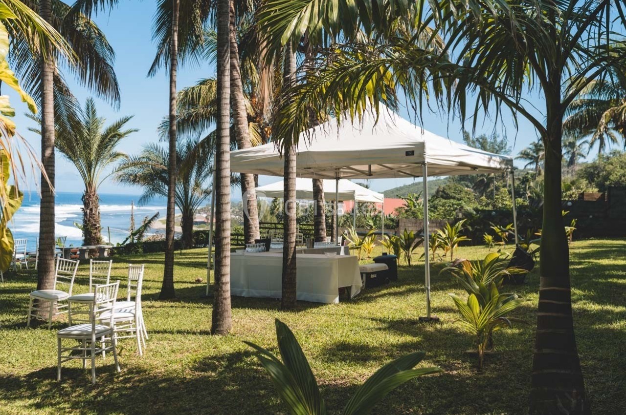 Tente blanche installée sur une pelouse entourée de palmiers, avec des chaises blanches et vue sur l'océan.