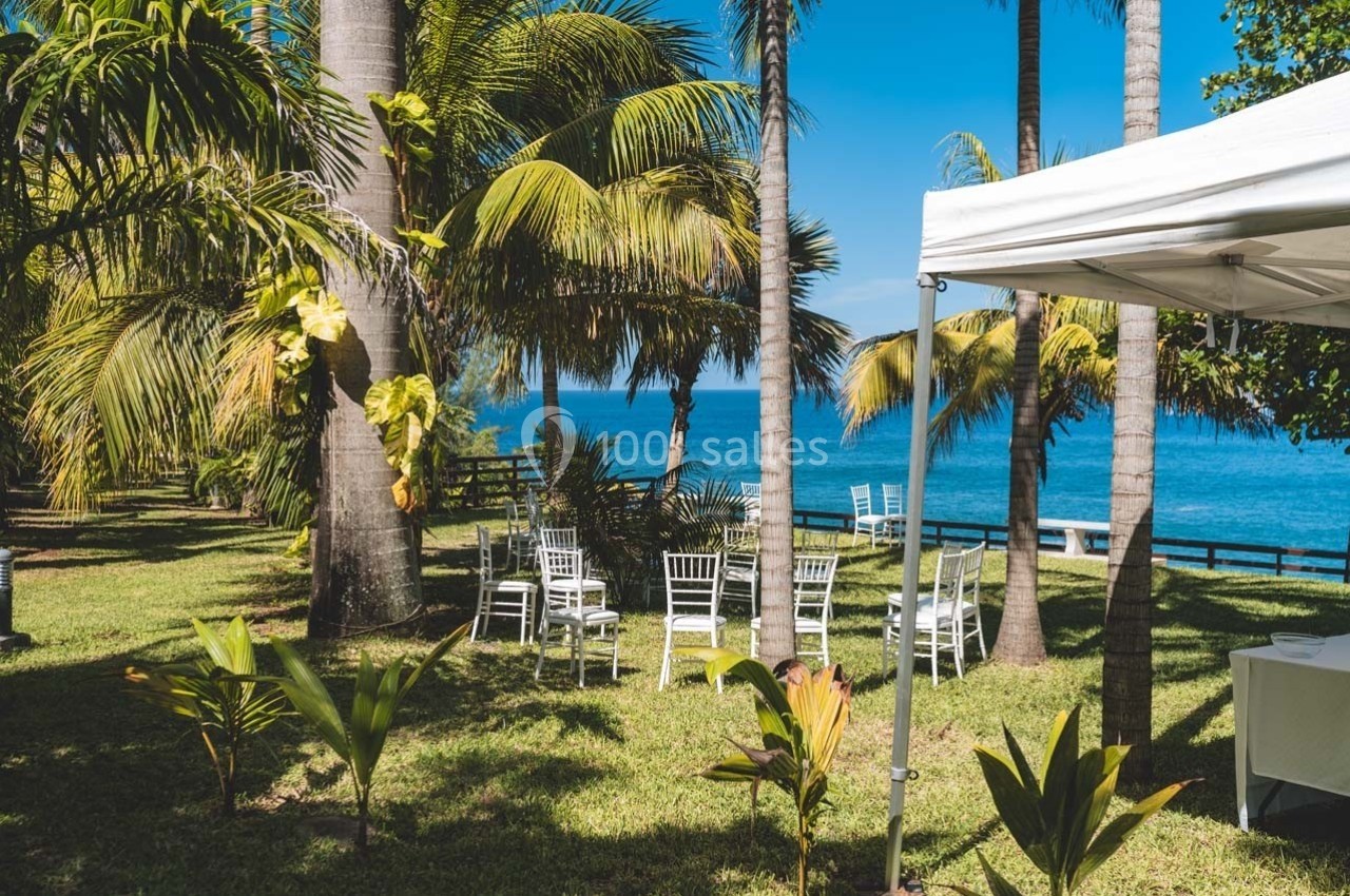 Chaises blanches disposées en cercle sur une pelouse entourée de palmiers, avec vue sur la mer sous un ciel bleu.