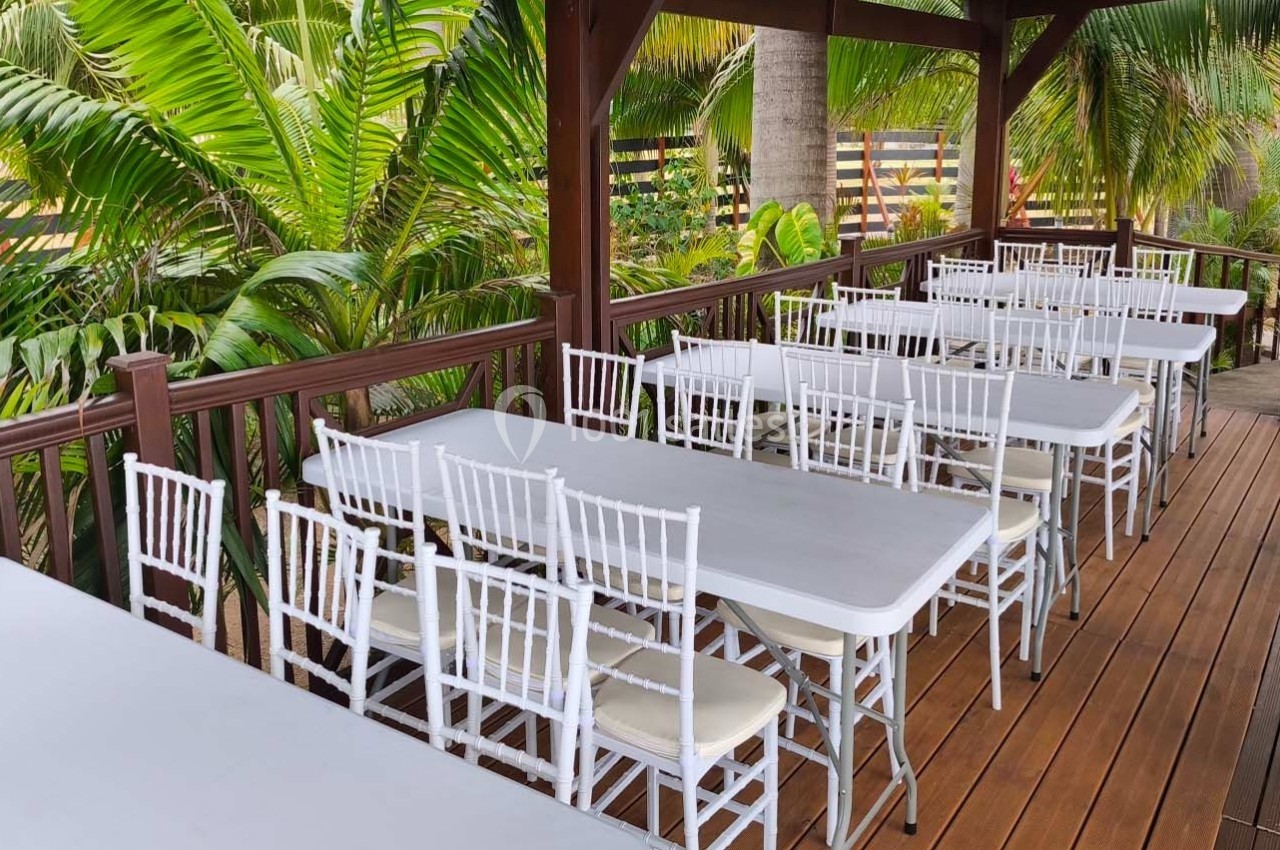 Terrasse en bois avec des tables rectangulaires blanches et des chaises assorties, entourée de palmiers.