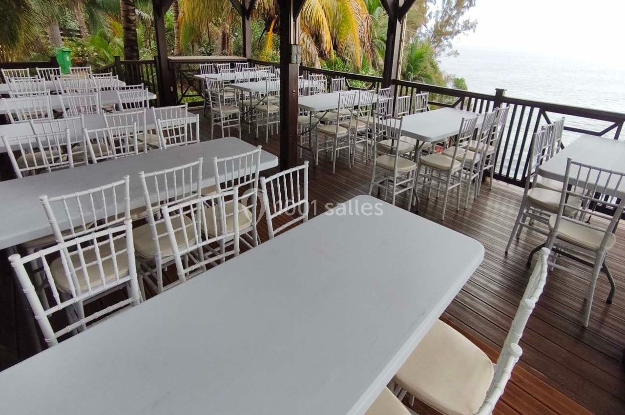 Terrasse en bois avec des tables et chaises blanches, offrant une vue sur la mer et entourée de végétation tropicale.