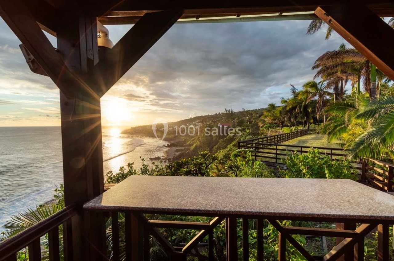 Vue sur un littoral tropical au coucher du soleil depuis une terrasse en bois avec une table en pierre.