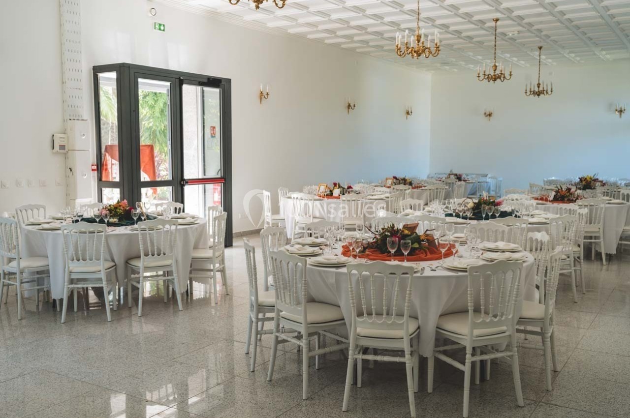 Salle de réception lumineuse avec des tables rondes dressées, décorées de centres de table floraux et chaises blanches.
