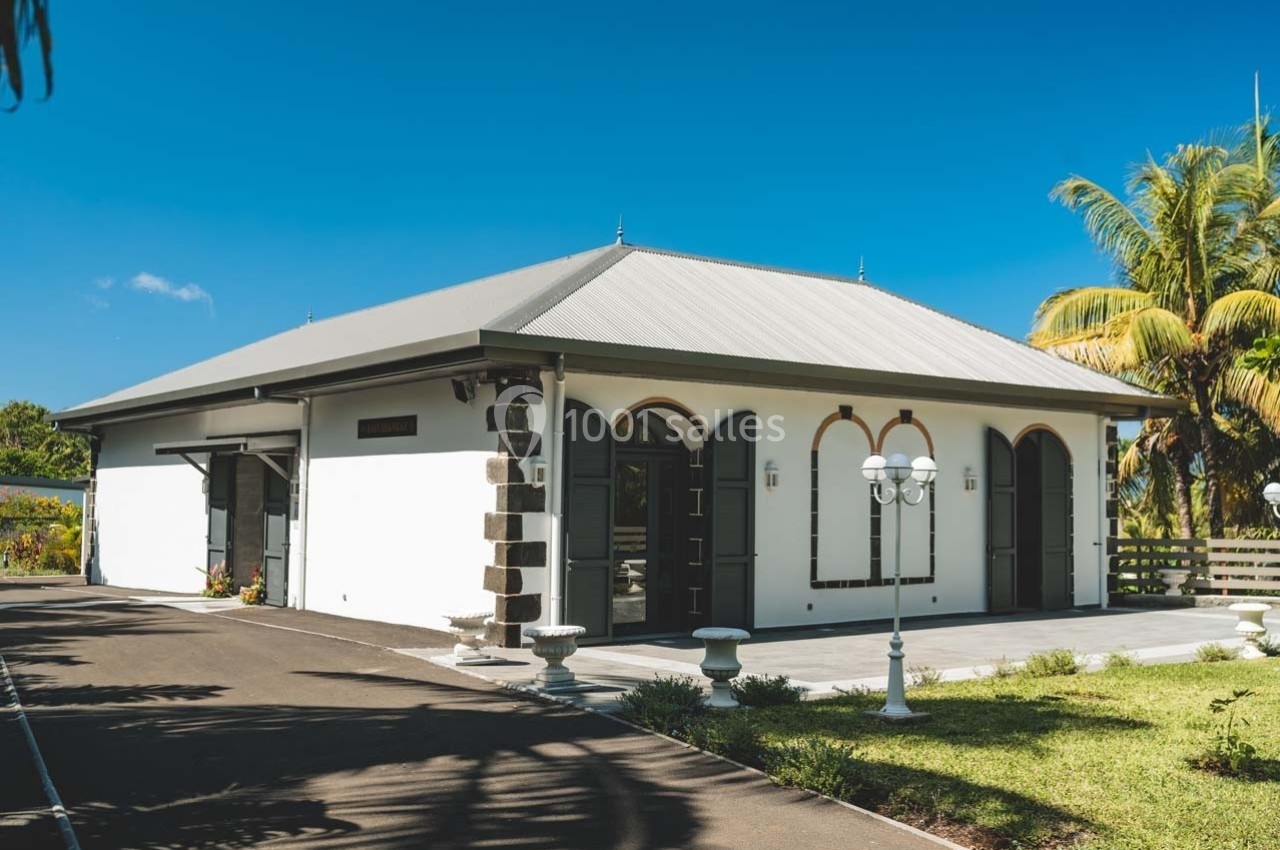Bâtiment blanc avec toit en métal gris, entouré de végétation tropicale et éclairé par un lampadaire.
