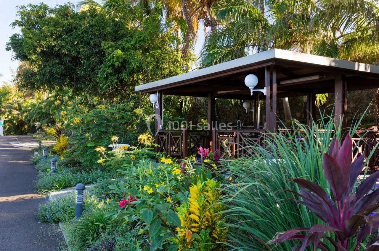 Abri en bois entouré de plantes tropicales et fleurs colorées, situé dans un jardin luxuriant.