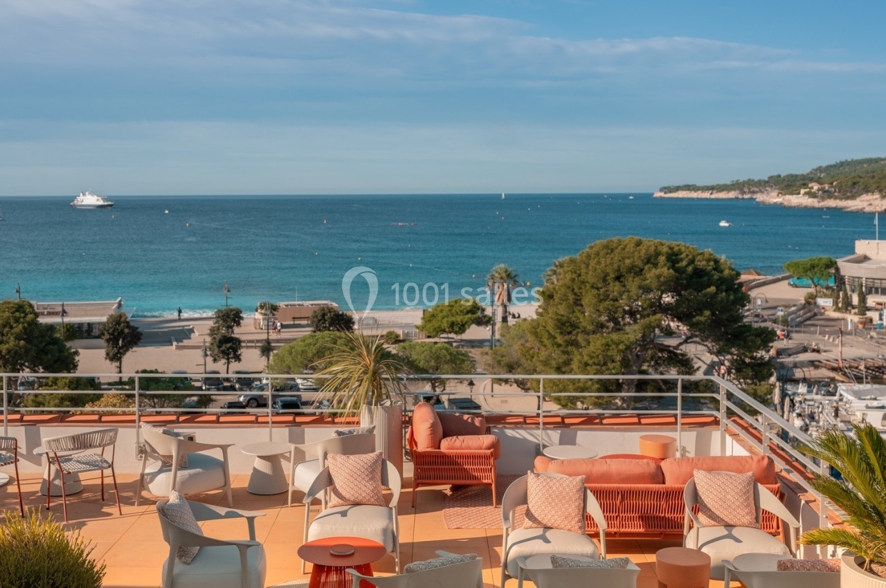 Terrasse aménagée avec mobilier coloré offrant une vue sur la mer, une plage, des arbres et un port au loin.