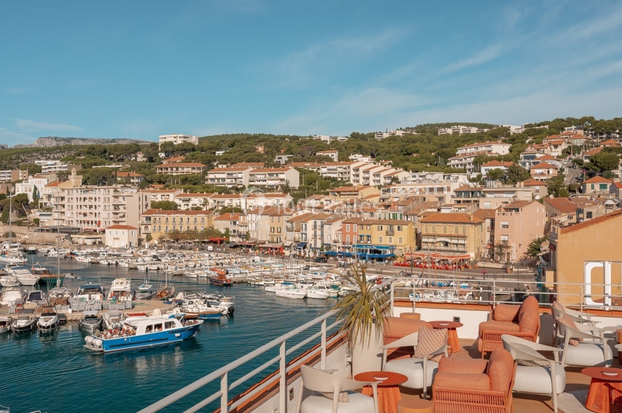 Vue d'une terrasse aménagée surplombant un port de plaisance entouré de bâtiments et de collines verdoyantes.