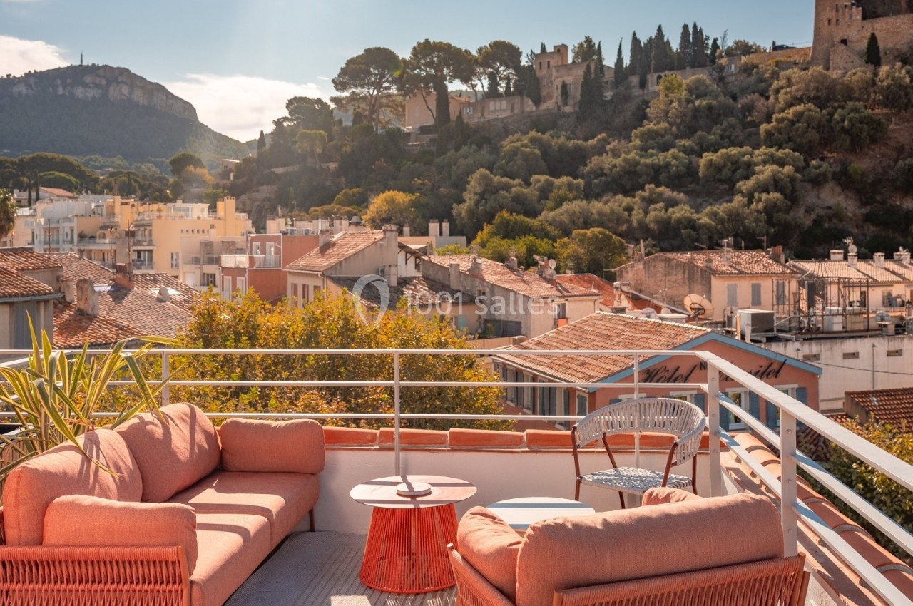 Terrasse aménagée avec des canapés et une table basse, offrant une vue sur des toits et une colline arborée avec des…