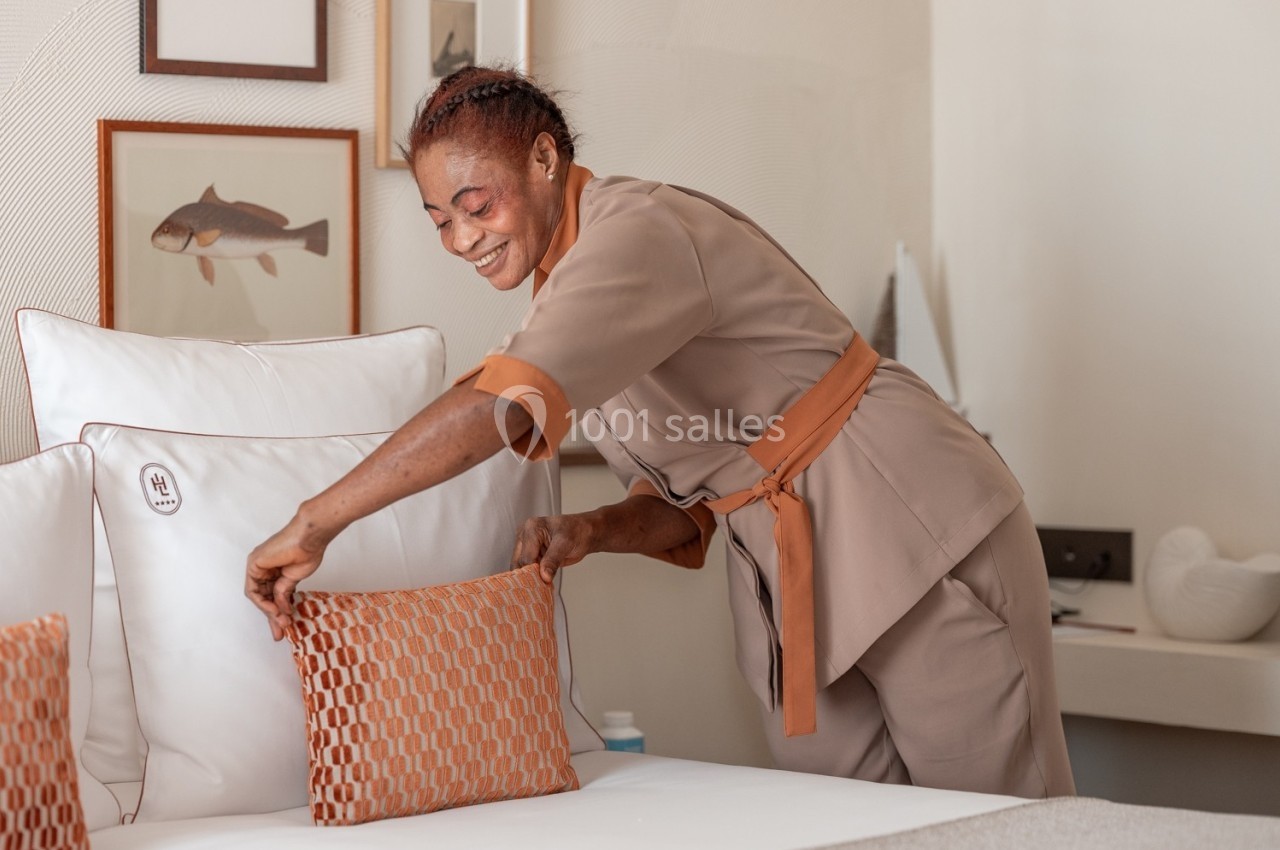 Une femme en uniforme arrange un coussin décoratif sur un lit dans une chambre lumineuse et élégante.