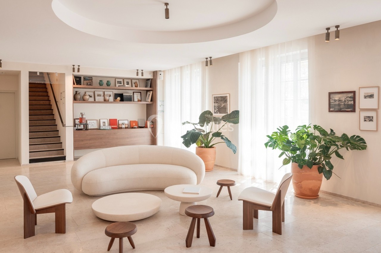 Salon lumineux avec canapé beige incurvé, chaises en bois, tables basses rondes et plantes en pot près de grandes fenêtres.