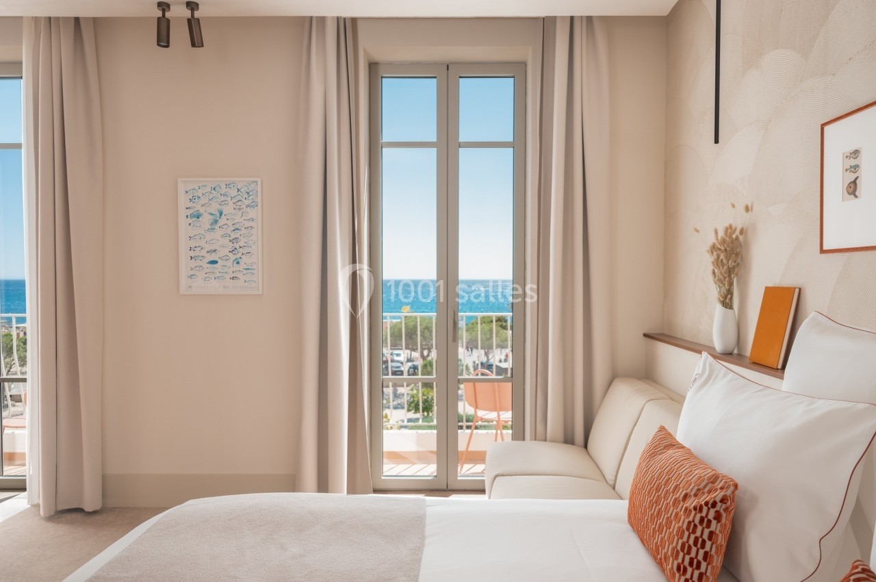 Chambre lumineuse avec lit, canapé, décoration murale et vue sur la mer à travers de grandes fenêtres.