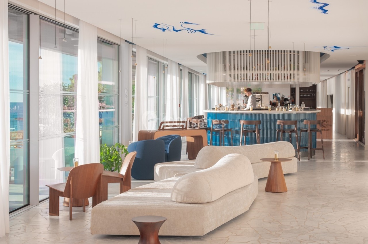 Salon lumineux avec mobilier moderne, bar en arrière-plan et grandes baies vitrées donnant sur l'extérieur.