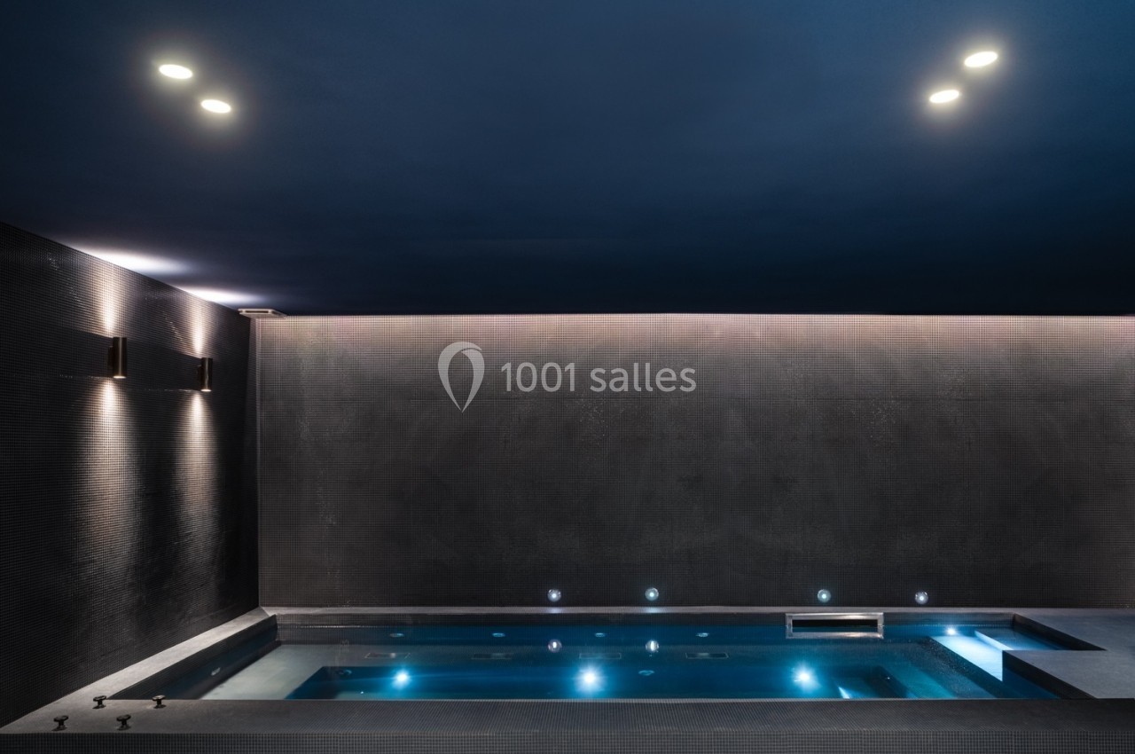 Piscine intérieure moderne éclairée par des lumières tamisées, avec un plafond sombre et des murs texturés.