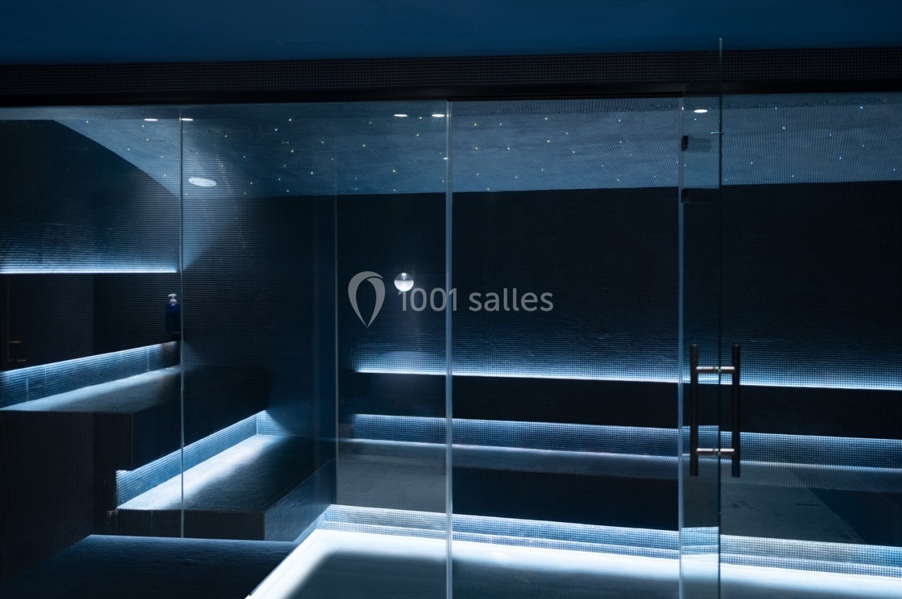 Salle de hammam moderne avec éclairage tamisé, murs sombres et bancs intégrés, vue à travers une porte vitrée.