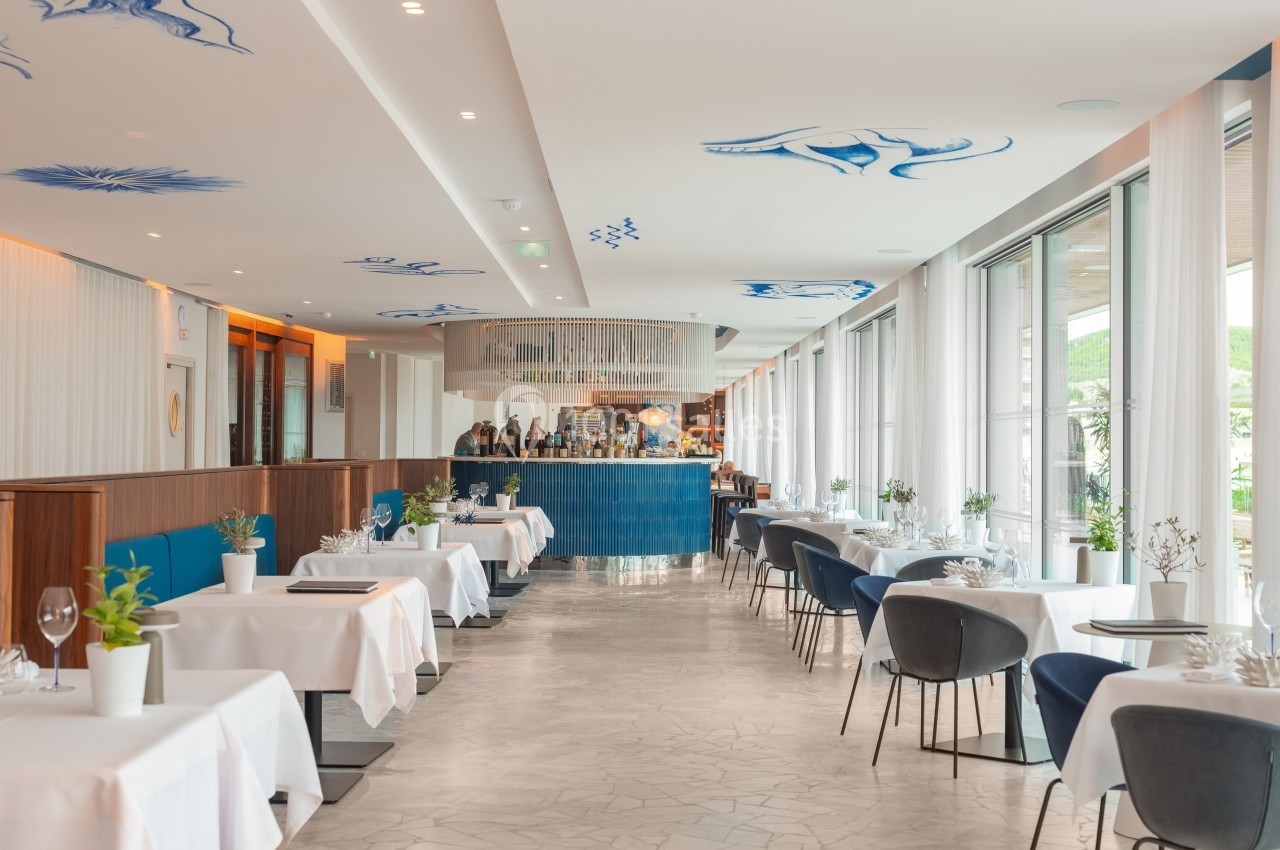 Salle de restaurant lumineuse avec tables dressées, chaises modernes et décoration bleue au plafond.