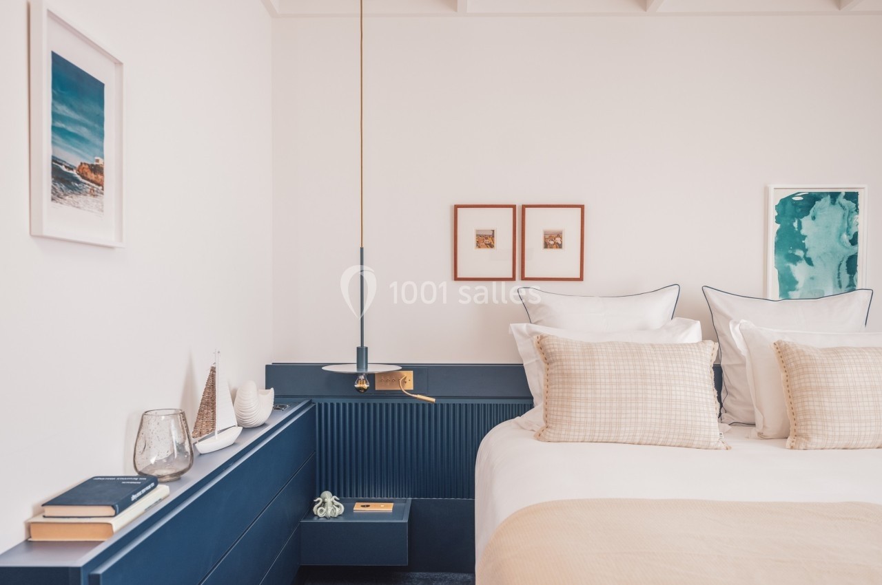 Chambre lumineuse avec lit double, table de chevet bleue, décor minimaliste et cadres accrochés au mur blanc.