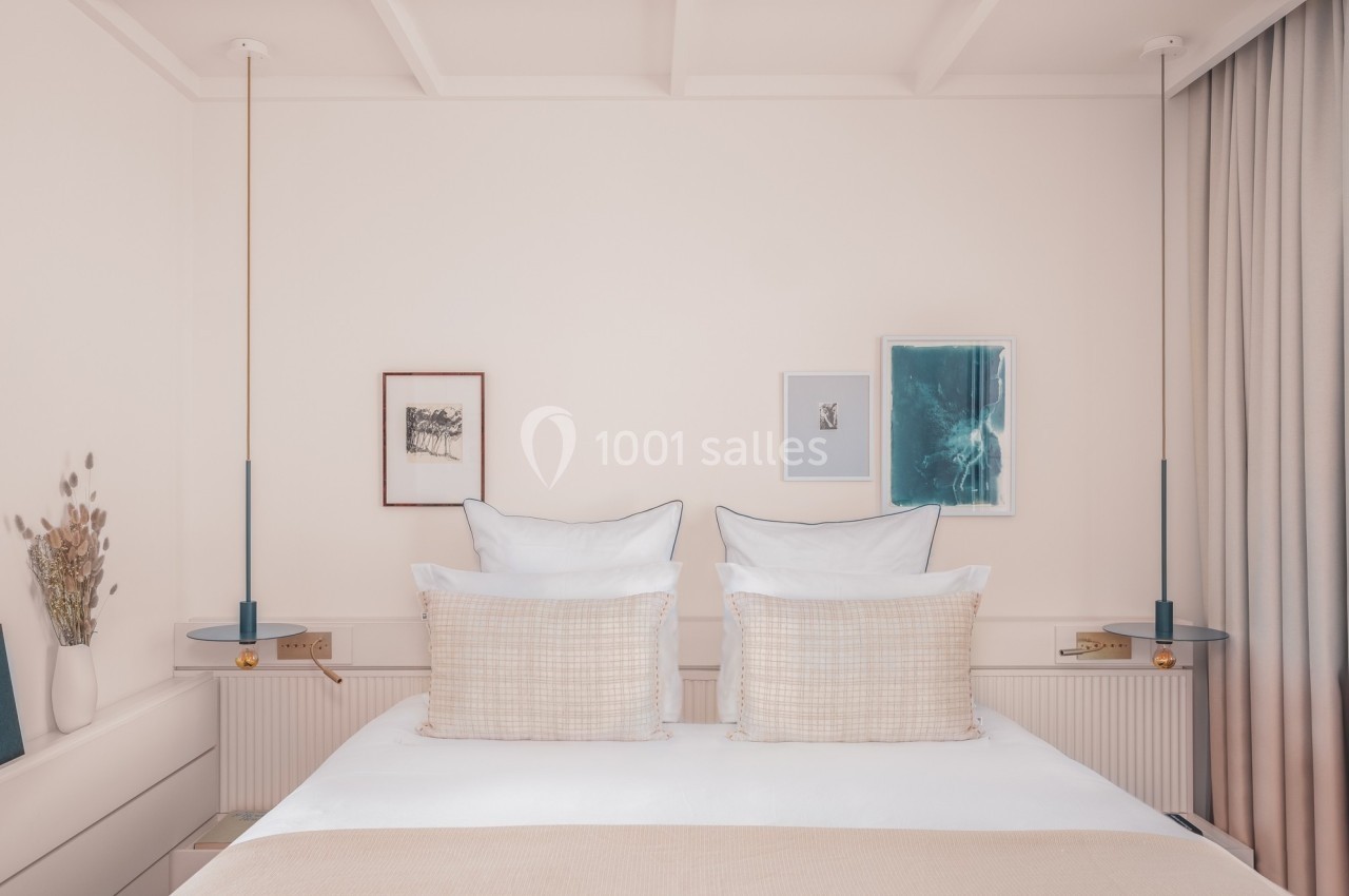 Lit double avec oreillers et coussins beiges, encadré par deux lampes suspendues et des tableaux sur un mur clair.