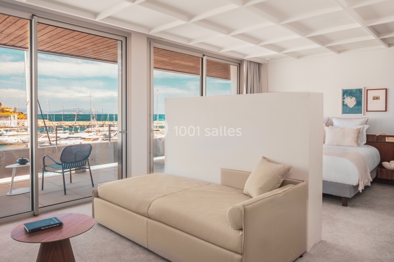 Vue d'une chambre lumineuse avec un canapé, un lit, et de grandes baies vitrées donnant sur un port de plaisance.