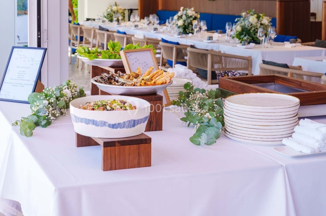 Buffet dressé sur une table blanche avec salades, pains et assiettes, décoré de fleurs et de verdure.