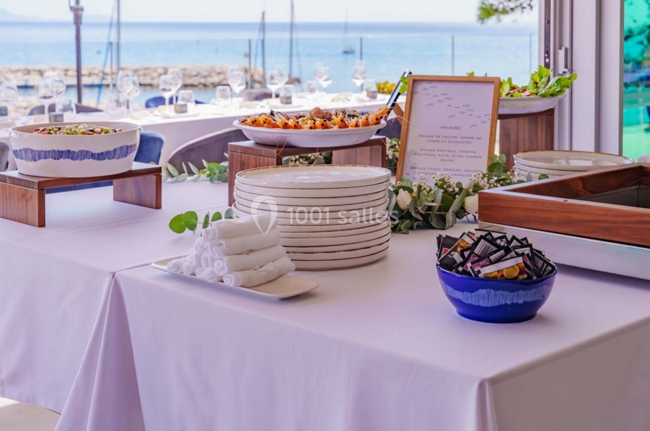 Table dressée avec vaisselle, serviettes pliées et plats variés, en arrière-plan une vue sur la mer et des tables…