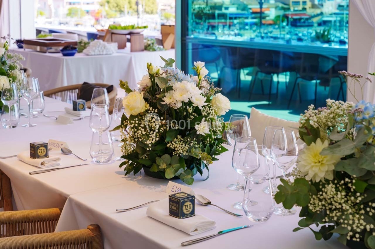 Table élégamment dressée avec nappes blanches, bouquets de fleurs et vaisselle raffinée, près d'une baie vitrée donnant sur…
