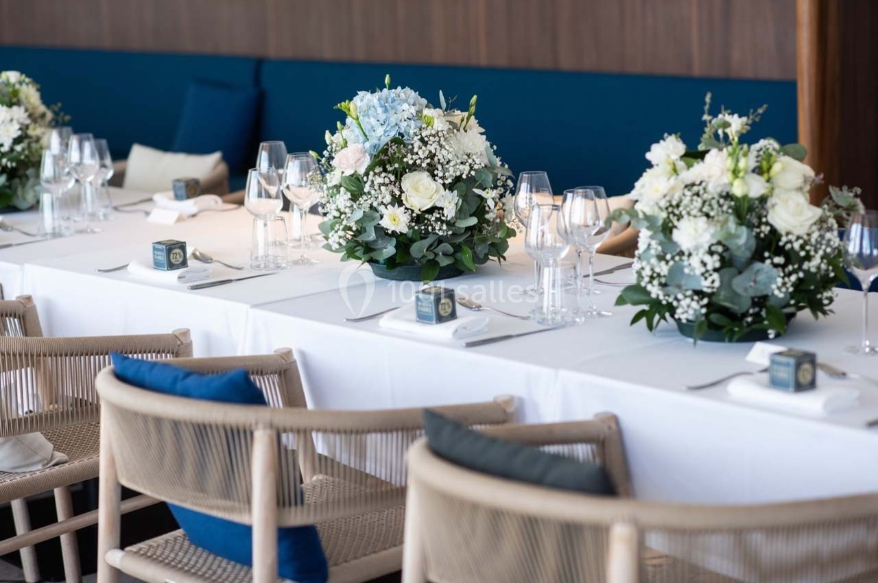 Table décorée pour un événement avec nappes blanches, bouquets de fleurs, verres à pied et chaises en bois clair.
