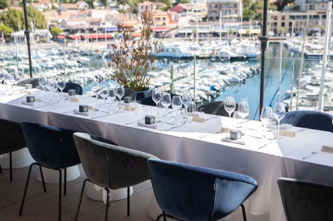 Table élégamment dressée avec nappes blanches et verres, donnant sur un port de plaisance en arrière-plan.