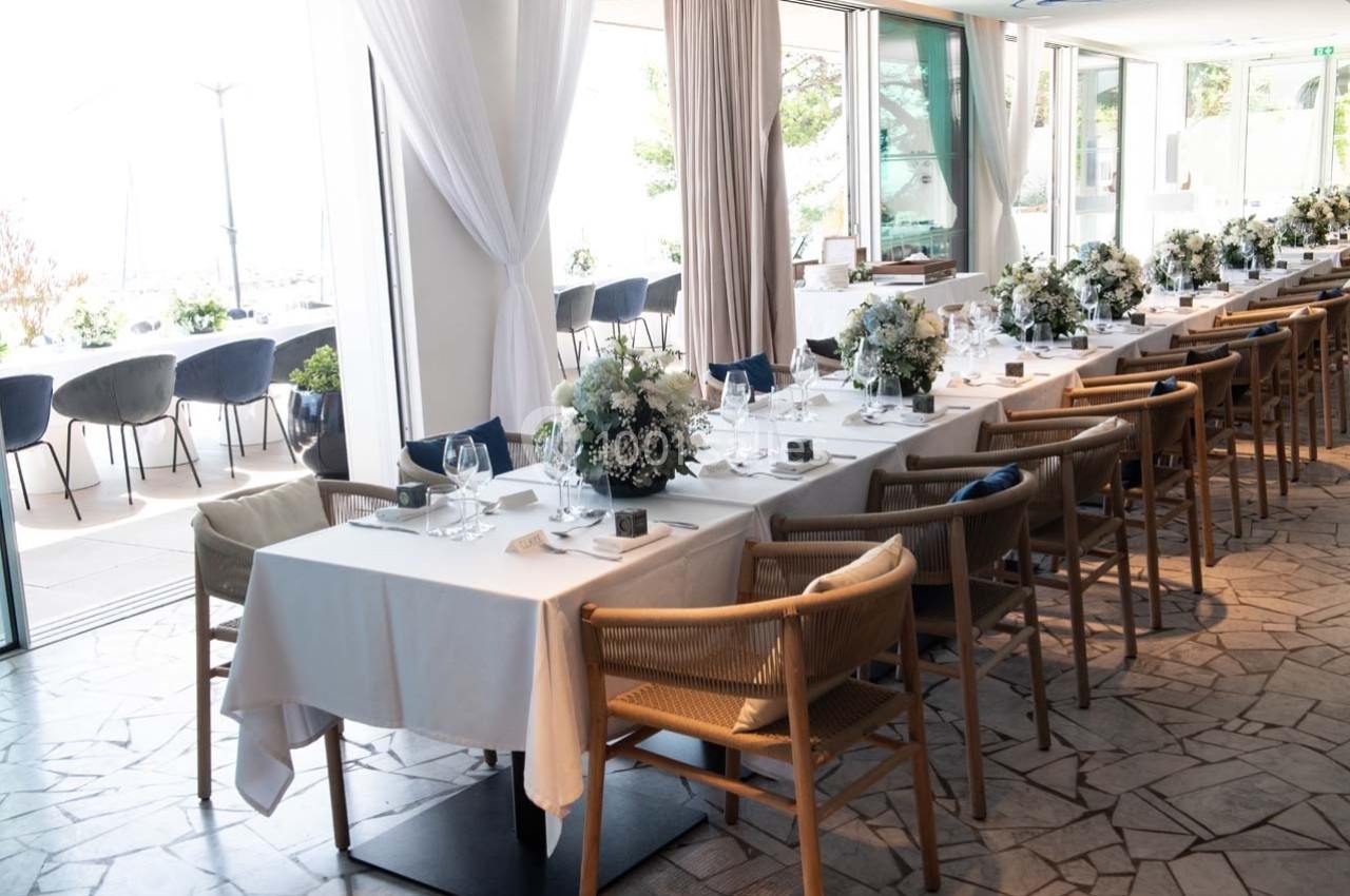 Salle de restaurant lumineuse avec de longues tables dressées, décorées de fleurs blanches et chaises en bois.