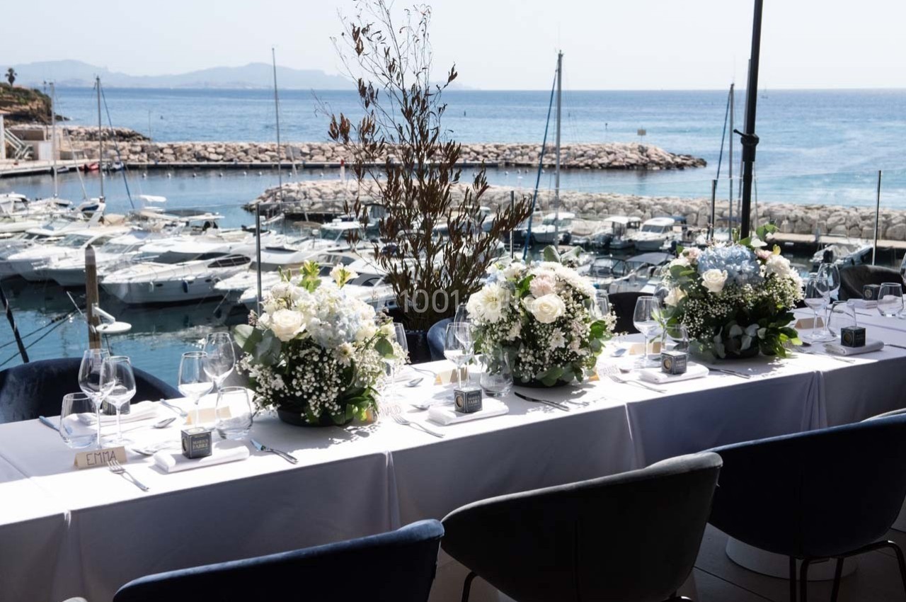 Table élégamment dressée avec des bouquets de fleurs, face à un port de plaisance et une vue sur la mer.