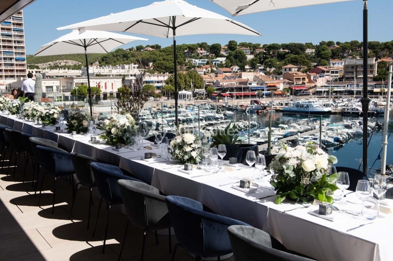 Tables élégamment dressées avec des fleurs blanches sur une terrasse surplombant un port de plaisance ensoleillé.