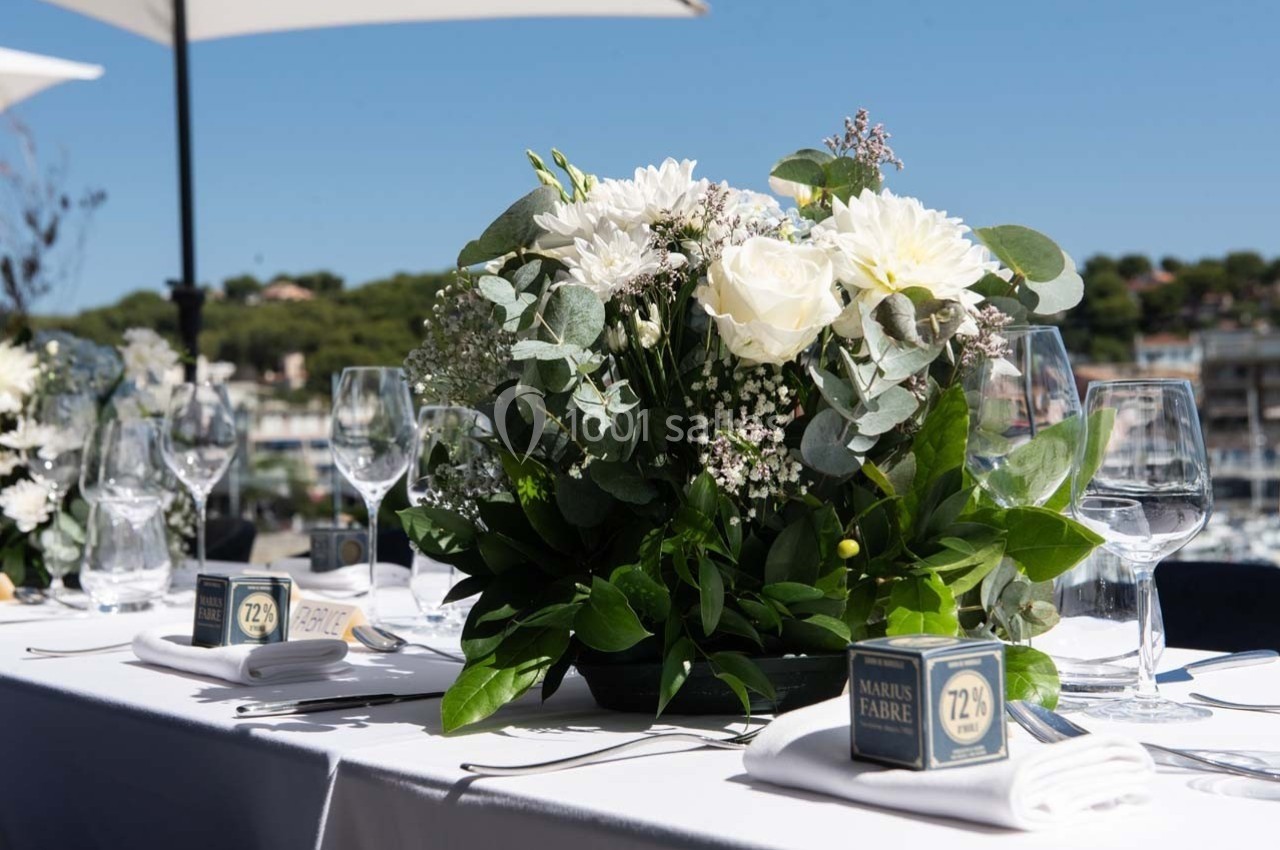 Centre de table floral avec roses blanches et feuillage, disposé sur une table dressée en extérieur sous des parasols.