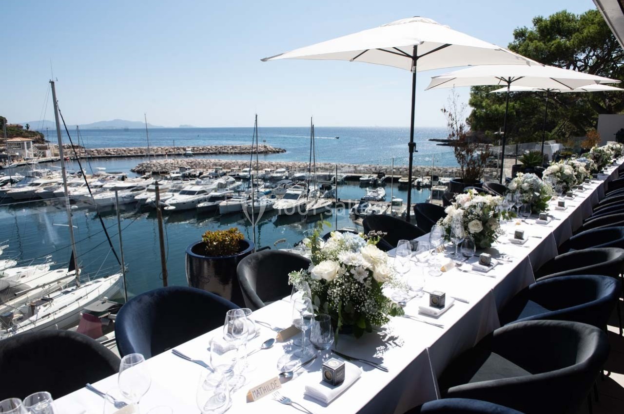 Grande table dressée avec des fleurs blanches en bord de mer, face à un port de plaisance sous un ciel ensoleillé.