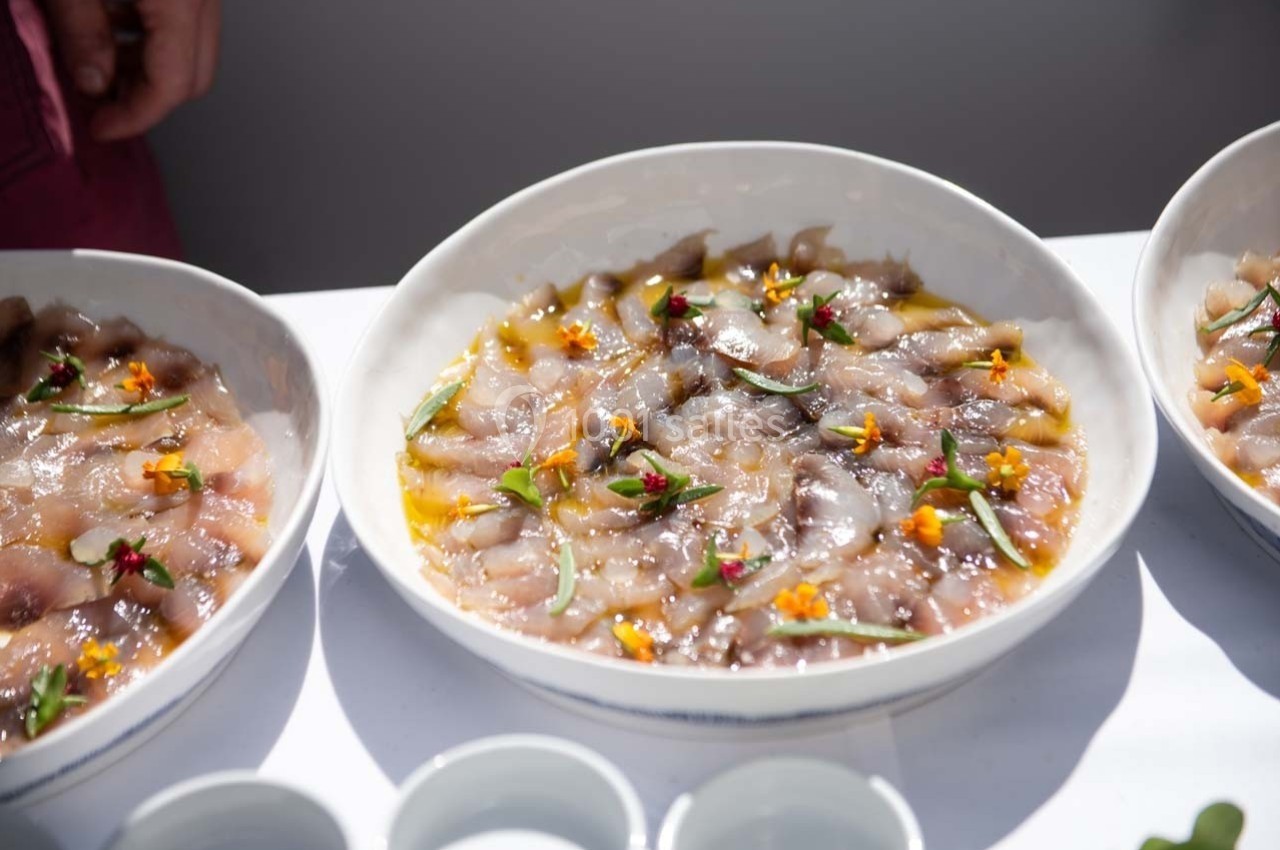 Assiette de carpaccio de poisson cru, décorée de fleurs comestibles et d'herbes fraîches, présentée sur une table blanche.