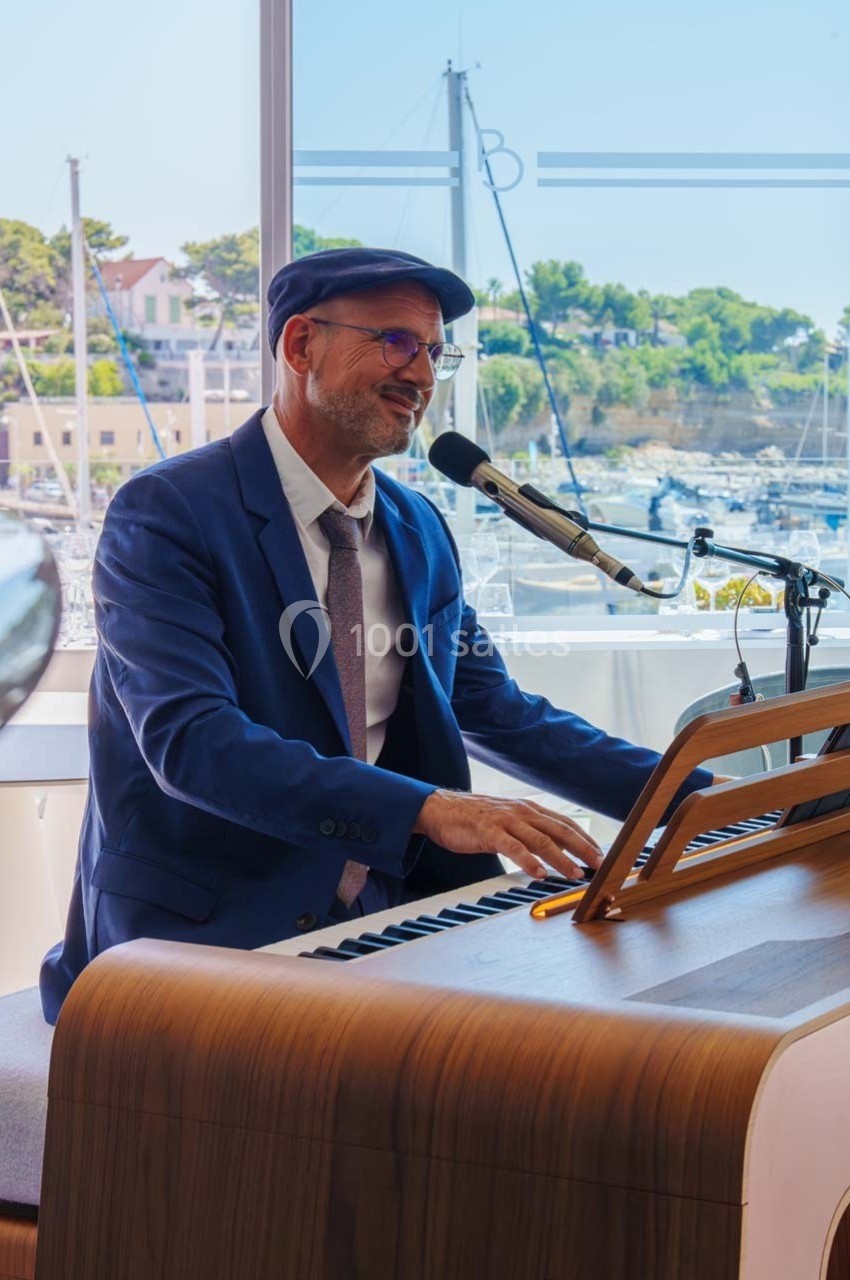Un homme en costume joue du piano et chante au micro, avec un port de plaisance visible en arrière-plan.