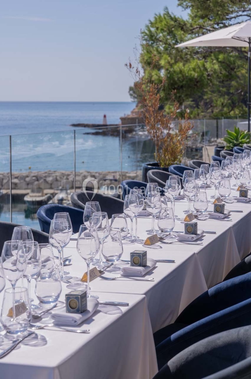 Table élégamment dressée en extérieur avec vue sur la mer, des verres et des assiettes alignés sous un ciel dégagé.