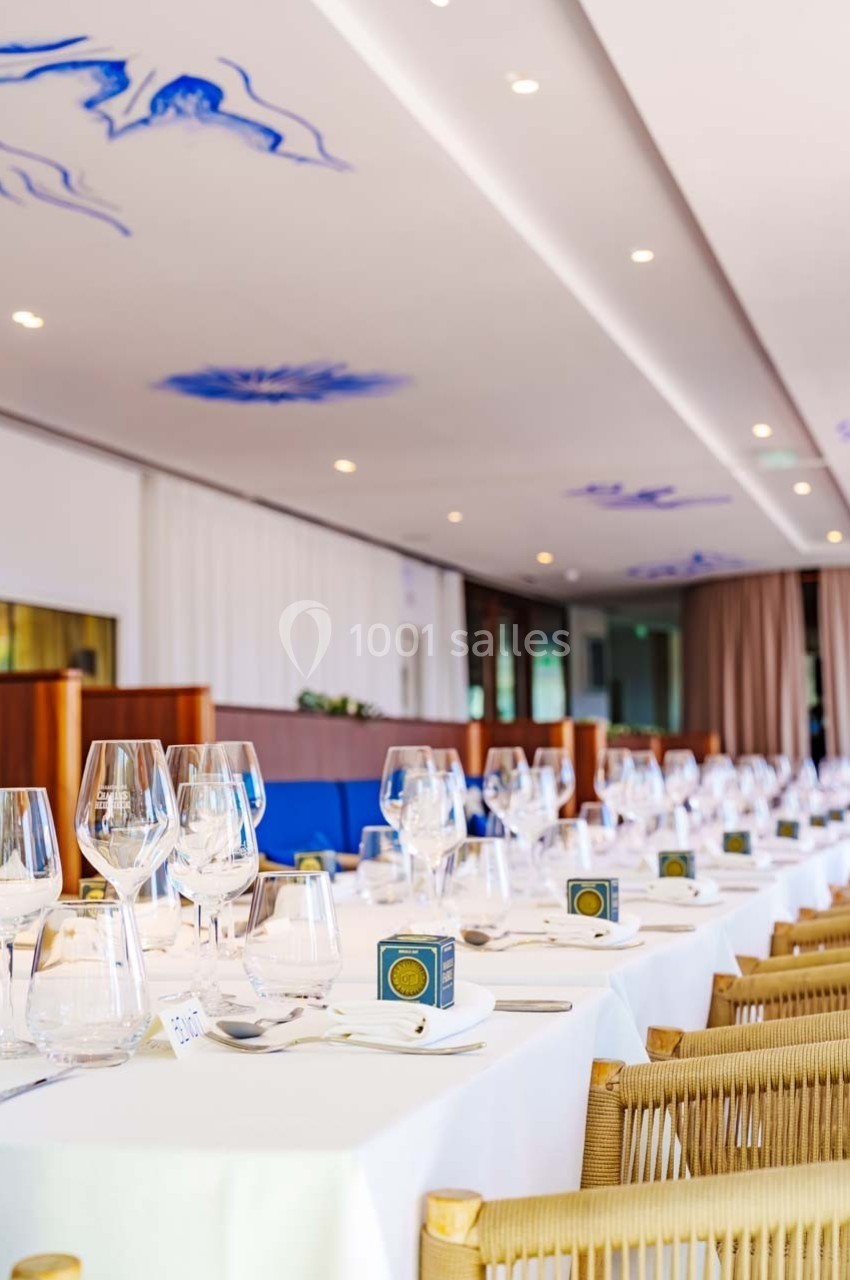 Salle de réception lumineuse avec tables dressées, nappes blanches, verres, couverts et décorations bleues au plafond.