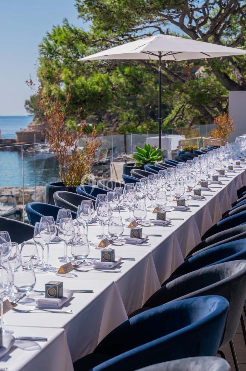 Table dressée en extérieur avec nappes blanches, verres et décorations, vue sur la mer et végétation environnante.