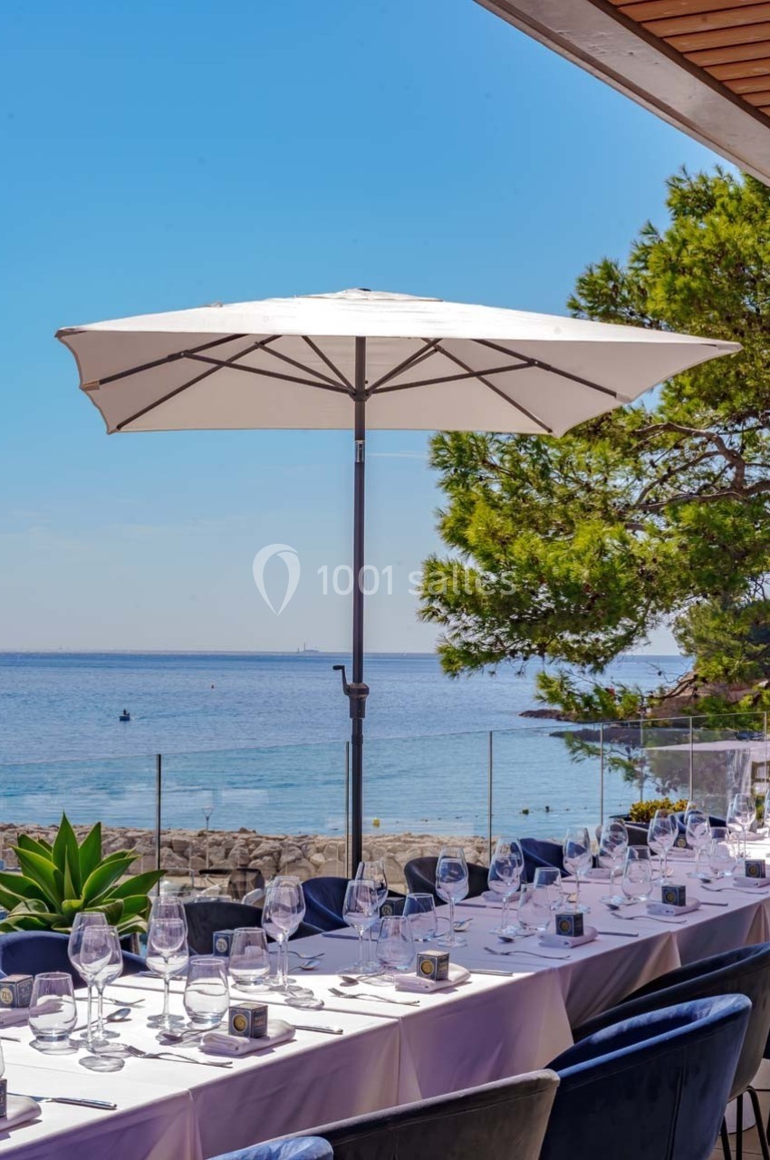 Table dressée en extérieur avec vue sur la mer, parasol blanc et végétation en arrière-plan.