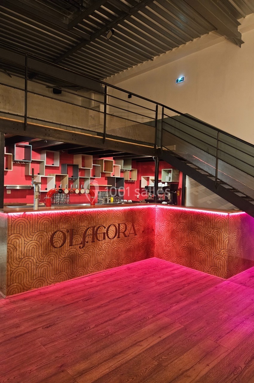 Comptoir de bar en bois avec éclairage rose, étagères géométriques rouges en arrière-plan et escalier métallique à droite.