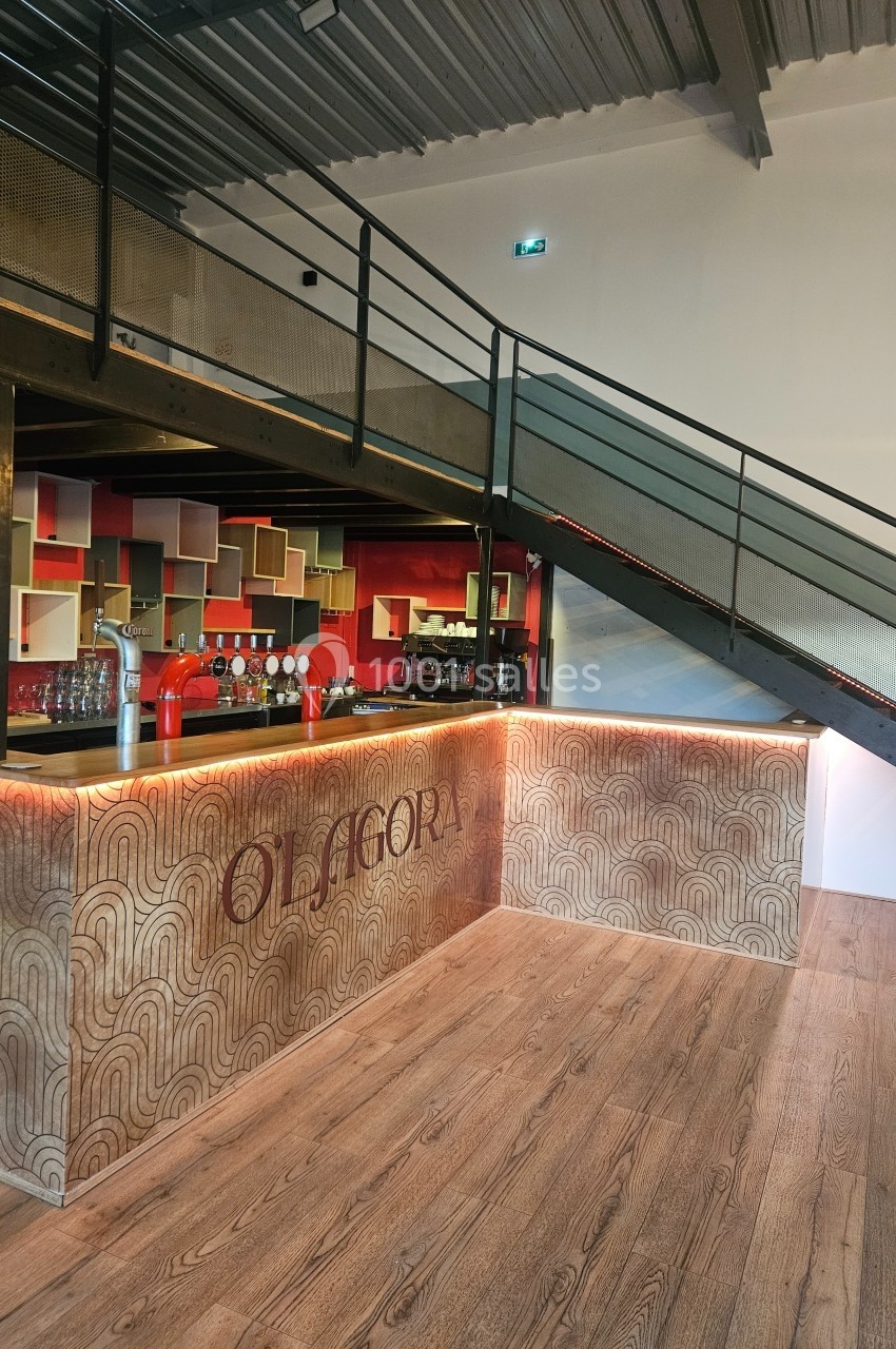 Comptoir de bar en bois décoré, situé dans un espace moderne avec un escalier métallique et des étagères colorées.