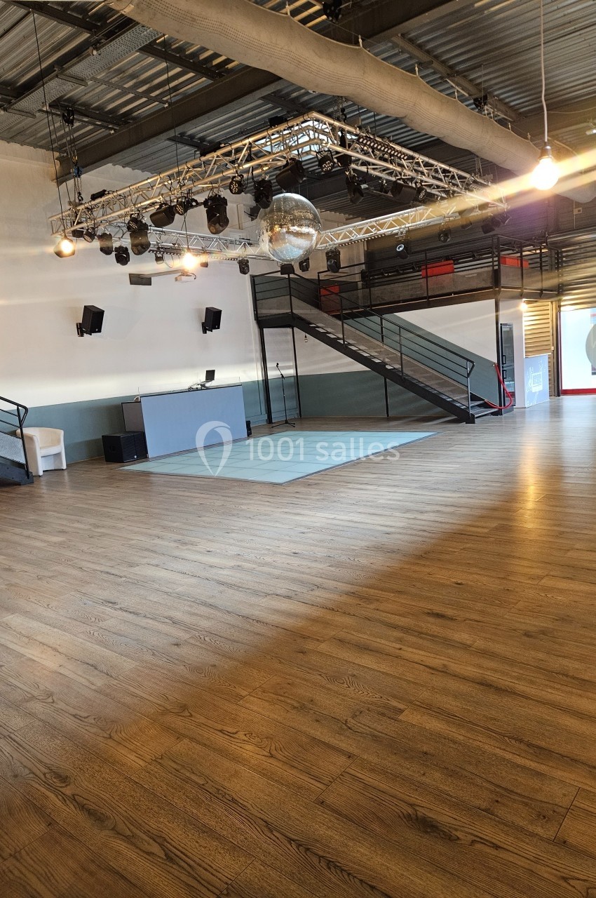 Salle spacieuse avec parquet, scène surélevée, éclairage suspendu et escalier menant à une mezzanine.