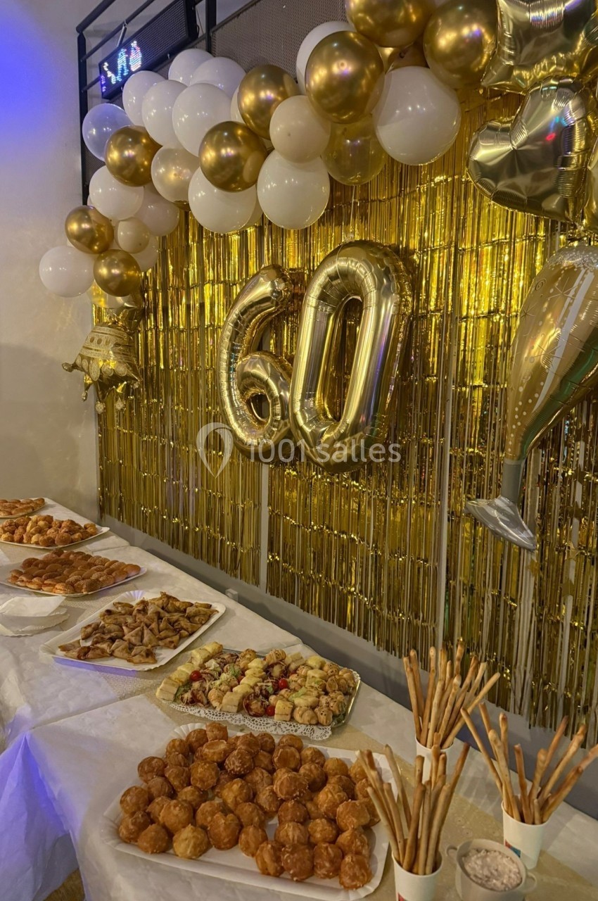 Décoration festive avec ballons dorés ’60’, guirlandes et buffet de pâtisseries sur des tables blanches.
