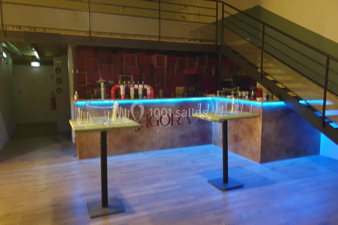 Bar avec éclairage bleu, tables hautes avec verres vides au premier plan, escalier métallique à droite.