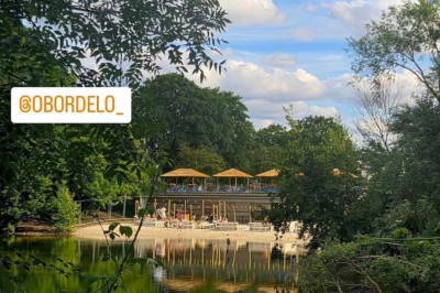 Deux personnes debout dans l'eau peu profonde d'un lac, tenant des verres, avec des arbres et un ciel bleu en arrière-plan.