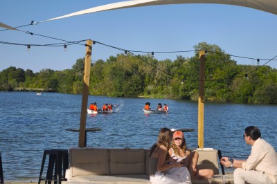 Deux personnes debout dans l'eau peu profonde d'un lac, tenant des verres, avec des arbres et un ciel bleu en arrière-plan.