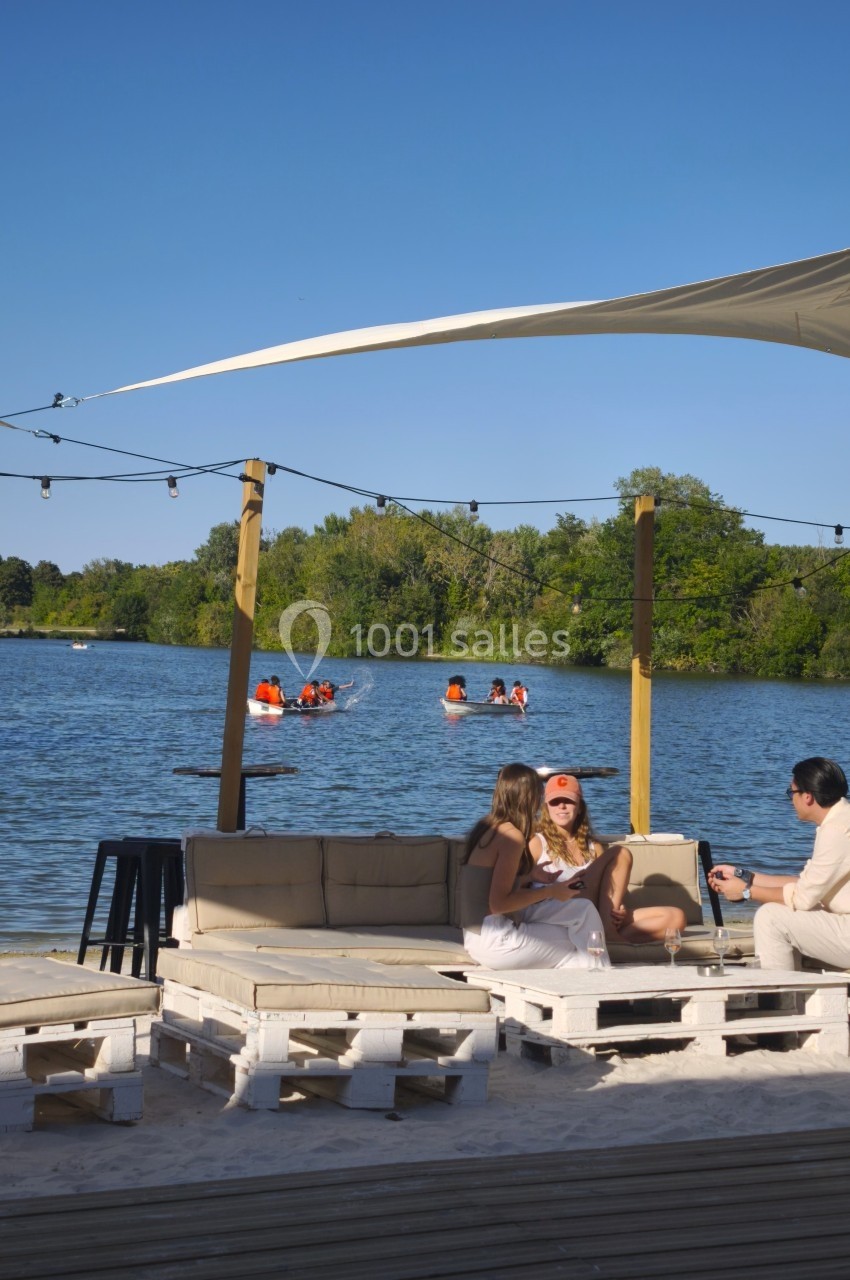 Des personnes discutent sur des canapés en palettes près d'un lac, tandis que d'autres font du canoë en arrière-plan.