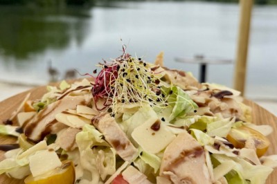 Deux personnes debout dans l'eau peu profonde d'un lac, tenant des verres, avec des arbres et un ciel bleu en arrière-plan.