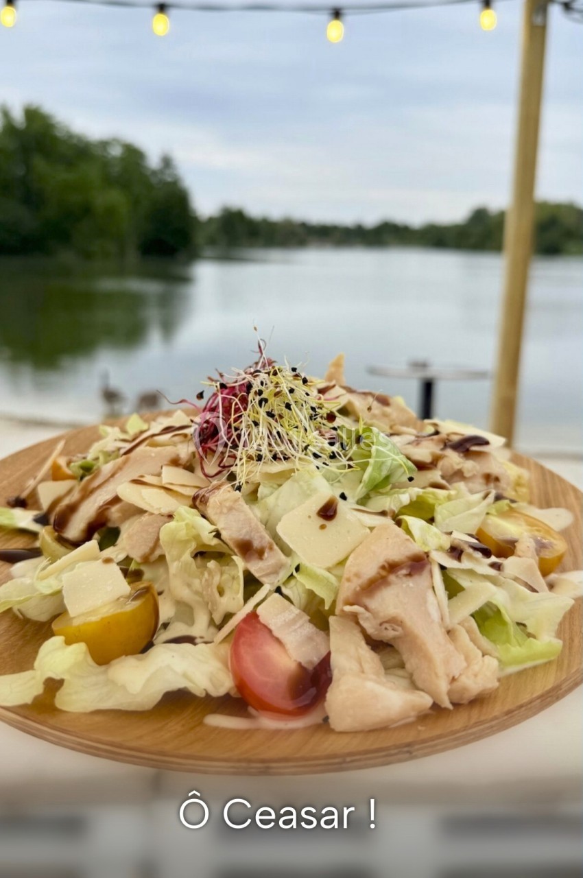 Assiette de salade César garnie de poulet, tomates cerises, parmesan et pousses, servie en terrasse au bord d'un lac.