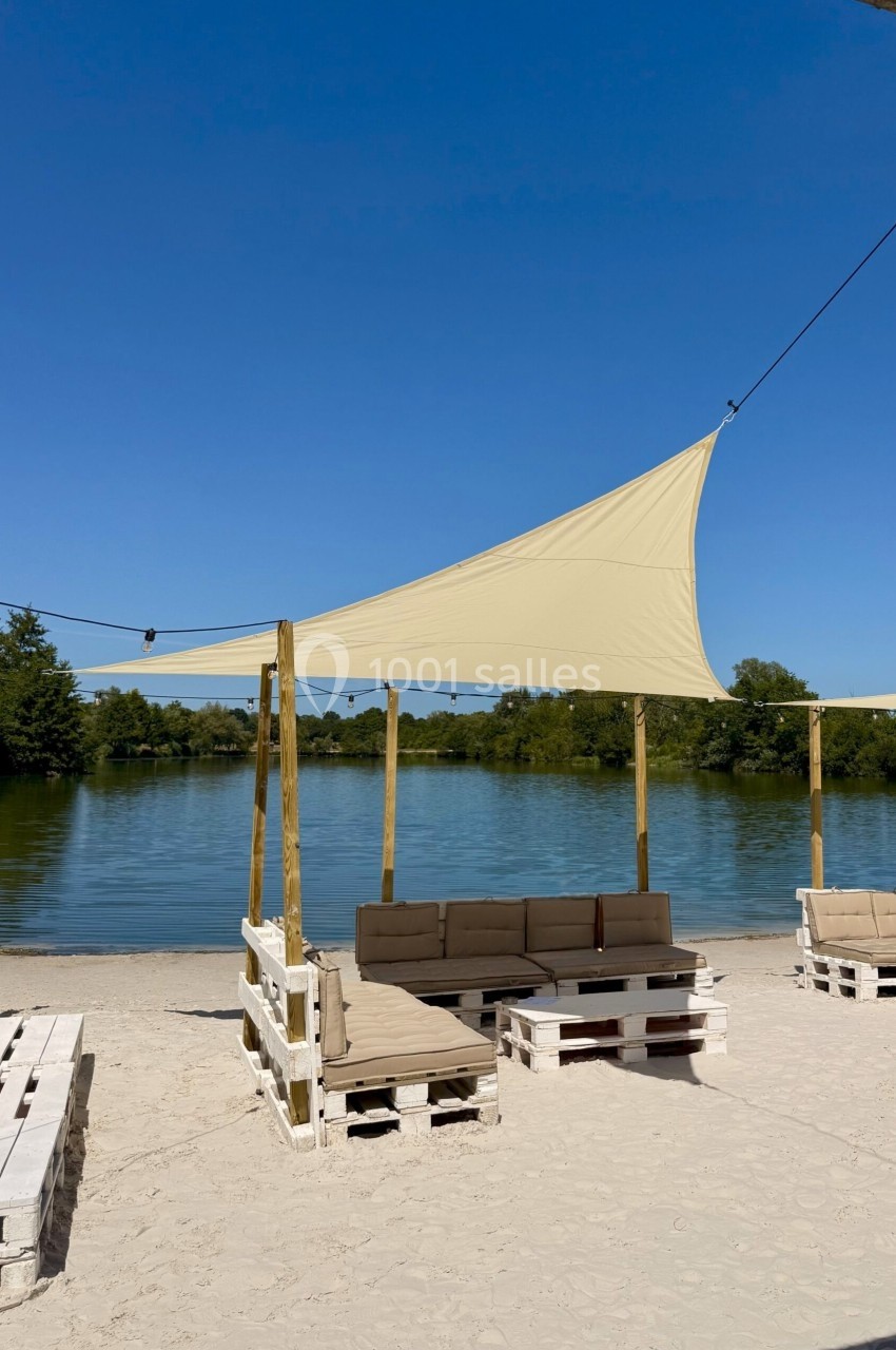 Canapé en palettes sous une voile d'ombrage beige, installé sur une plage au bord d'un lac entouré de verdure.