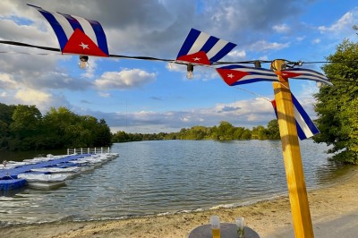 Deux personnes debout dans l'eau peu profonde d'un lac, tenant des verres, avec des arbres et un ciel bleu en arrière-plan.